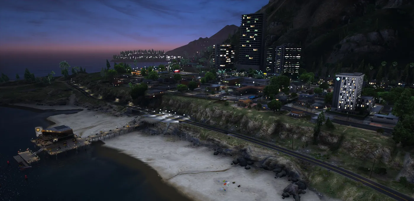 Paleto Bay at dusk from above the beach, with a lit café on the end of the pier, the town behind, a tall hospital on the right and apartment towers against the mountain